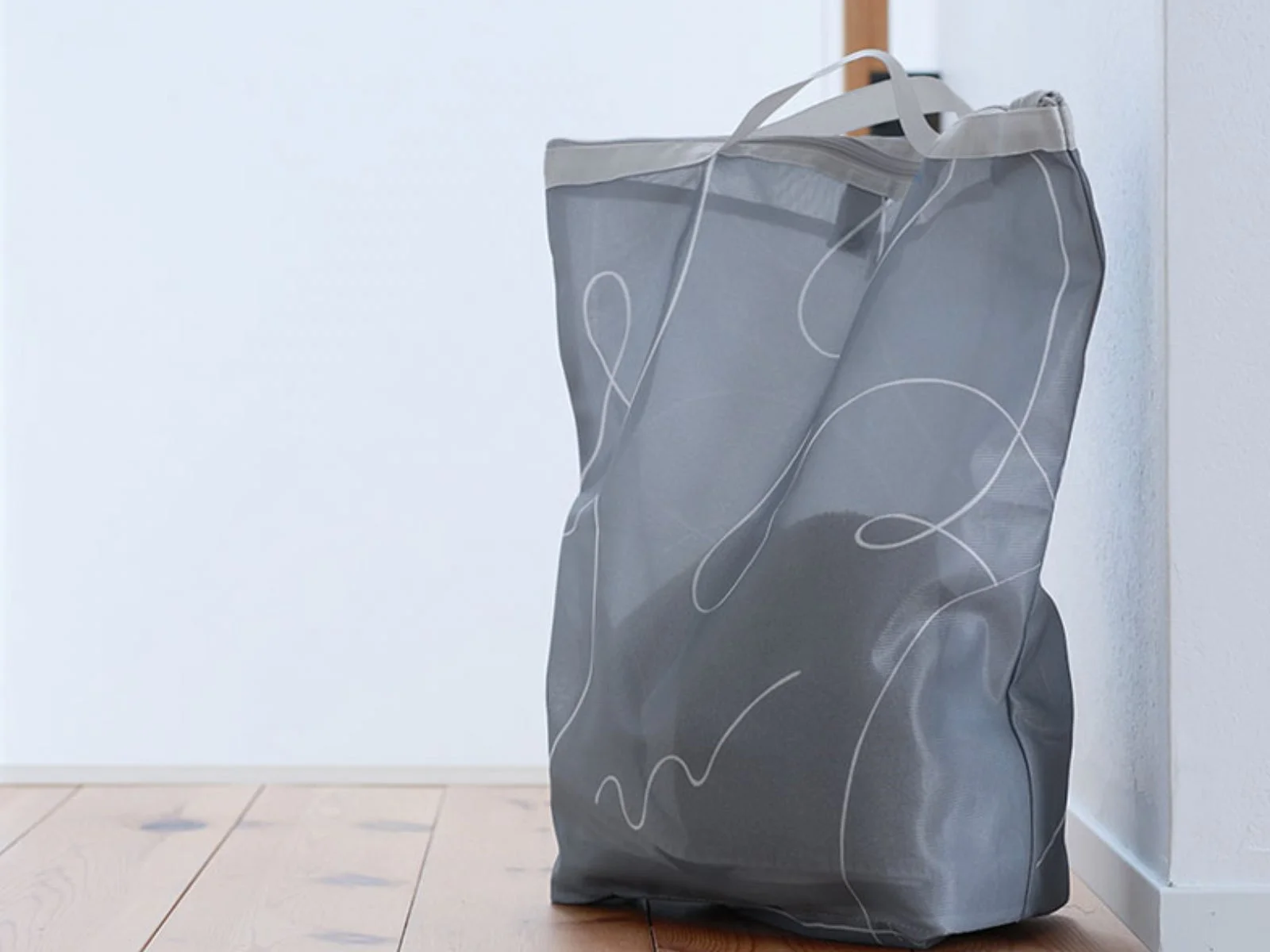 白い壁と木製の床を背景に、グレーの半透明なメッシュ製ランドリーバッグが置かれています。バッグには白い抽象的な線模様が描かれており、上部には持ち手とファスナーが見えます。シンプルで機能的なデザインです。