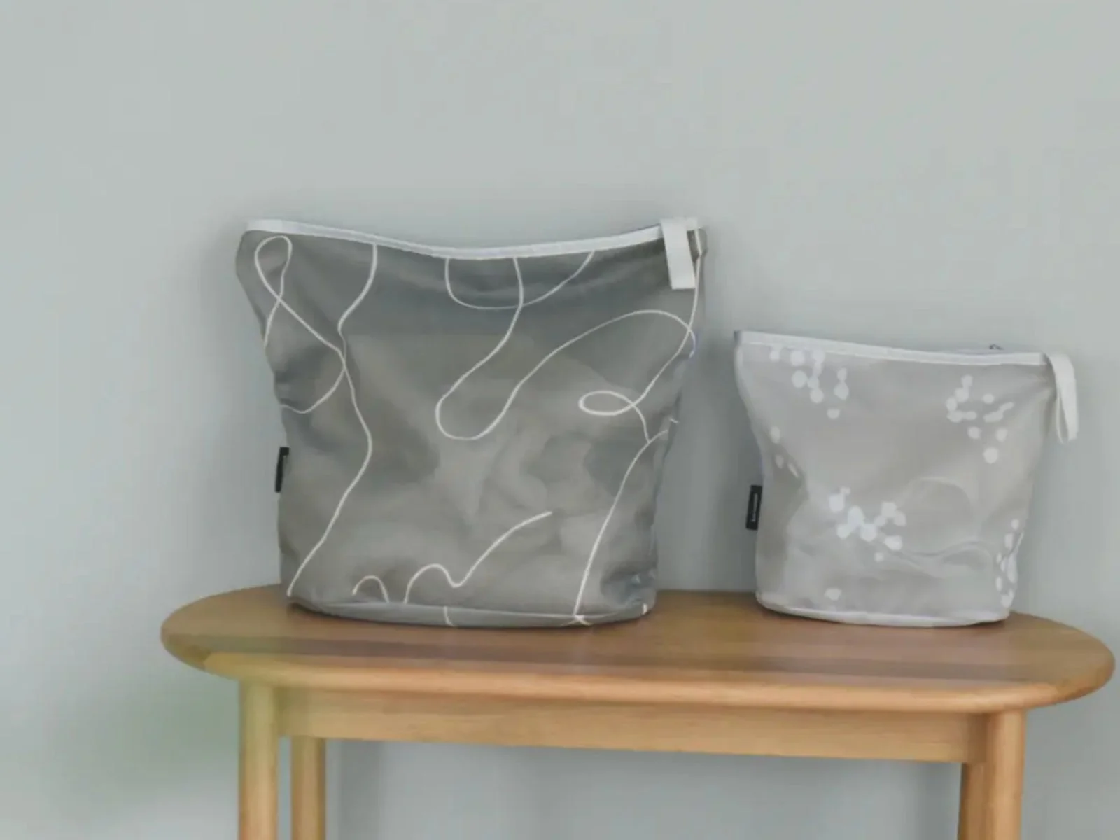 木製テーブルの上に、大小2つの半透明なメッシュポーチが置かれています。大きい方は曲線柄、小さい方はドット柄で、グレーを基調としたシンプルなデザインが特徴です。